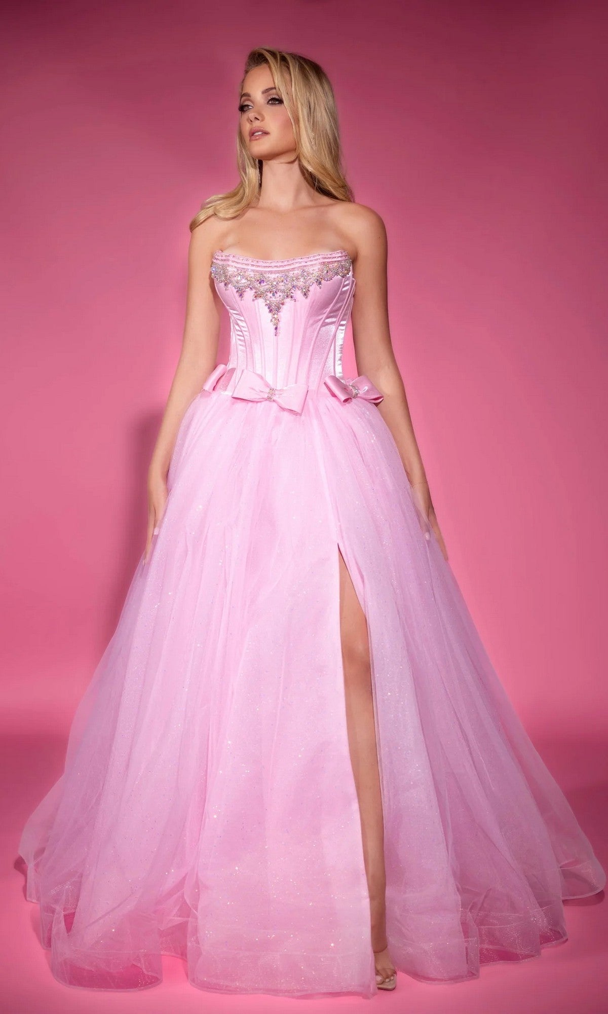 A woman poses against a pink background wearing the Portia and Scarlett PS25336, a long pink formal dress with a strapless corset bodice, beaded details, high slit, and glitter tulle accents.